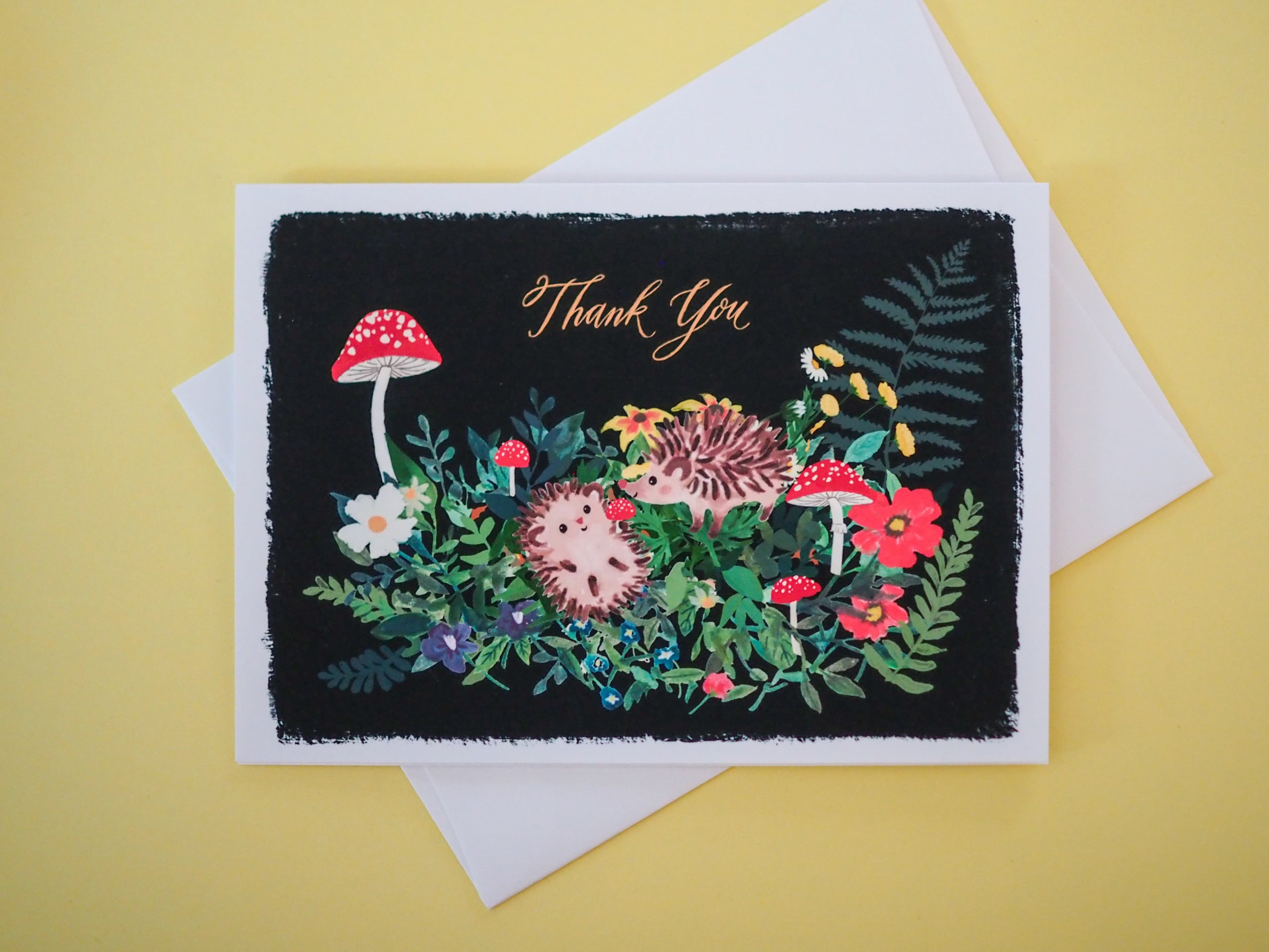 A folded thank you card featuring an illustration of a hedgehog, mushrooms, and flowers on a black background, with the words 'Thank You' displayed at the top.