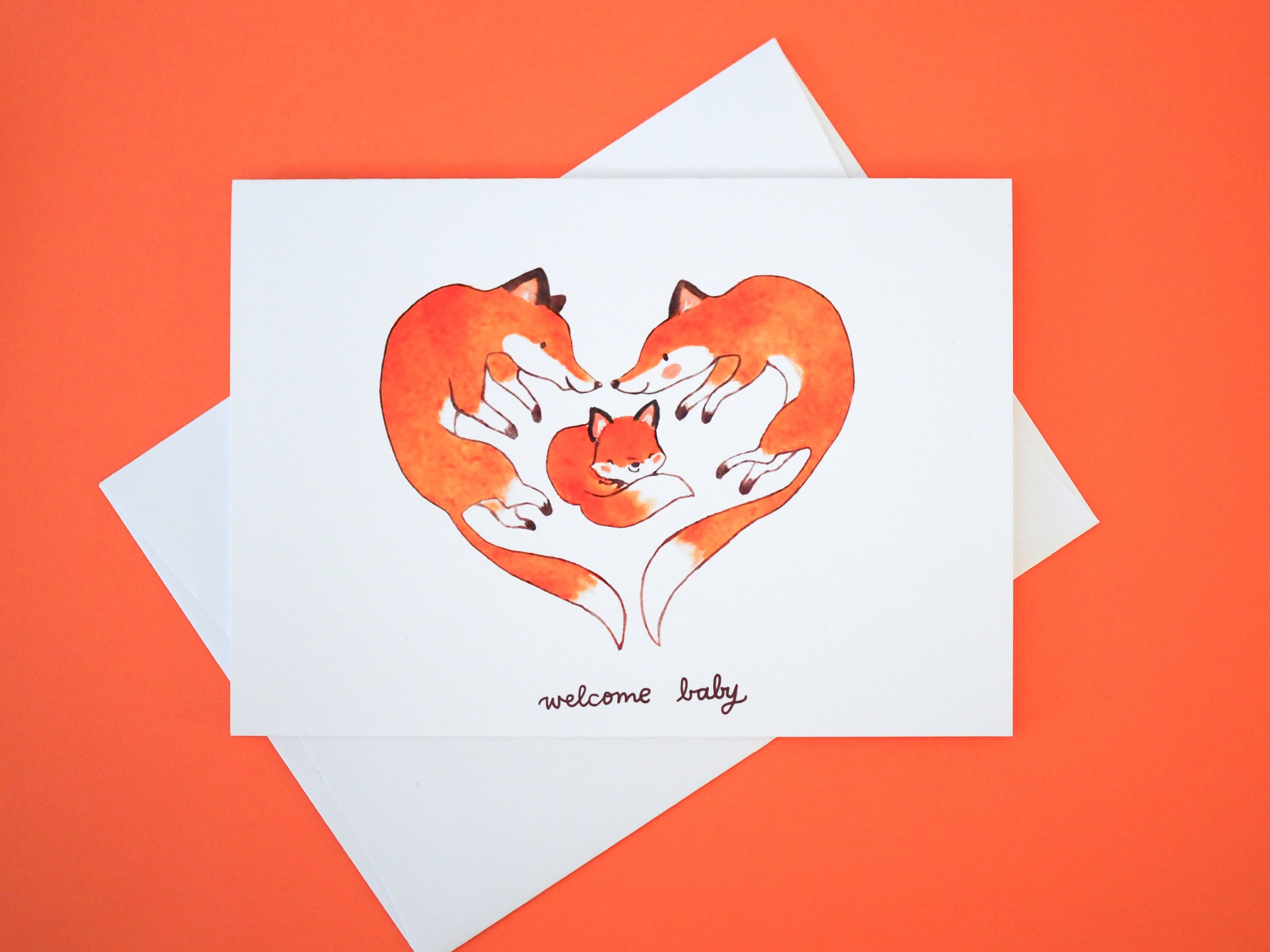 A greeting card with an illustration of two foxes forming a heart shape around a baby fox and the text 'welcome baby' in the center on an orange background.